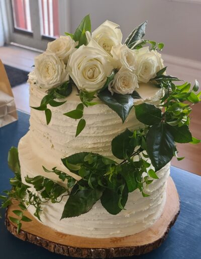 A white, two tier wedding cake topped with real white roses and greenery that wraps around the cake. The cake was created by Simply Baked Catering Inc. in Winchester, Ontario.
